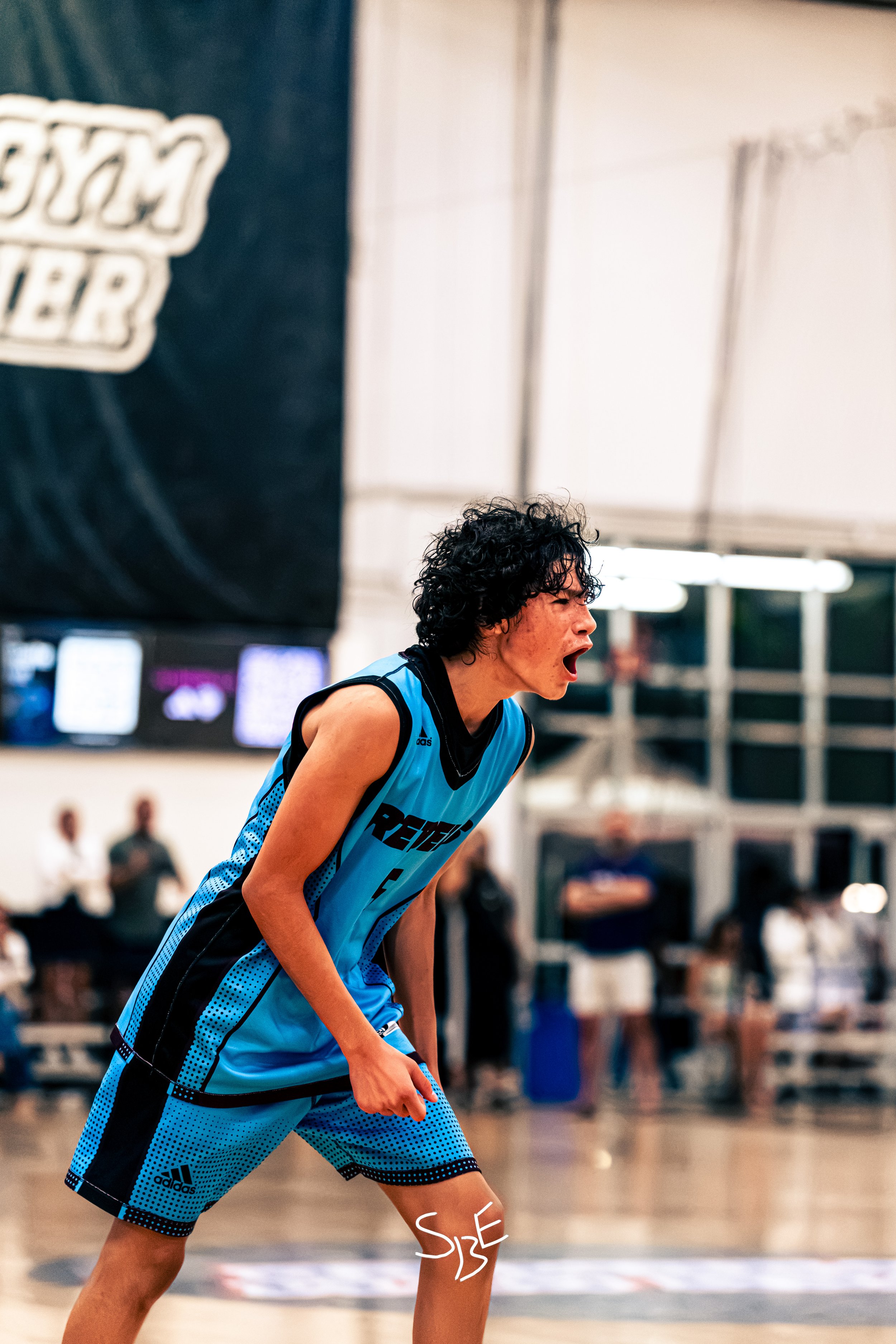 A young male basketball player with curly black hair, wearing a blue and black Adidas basketball uniform, appears to be shouting or expressing excitement on the court during a game.