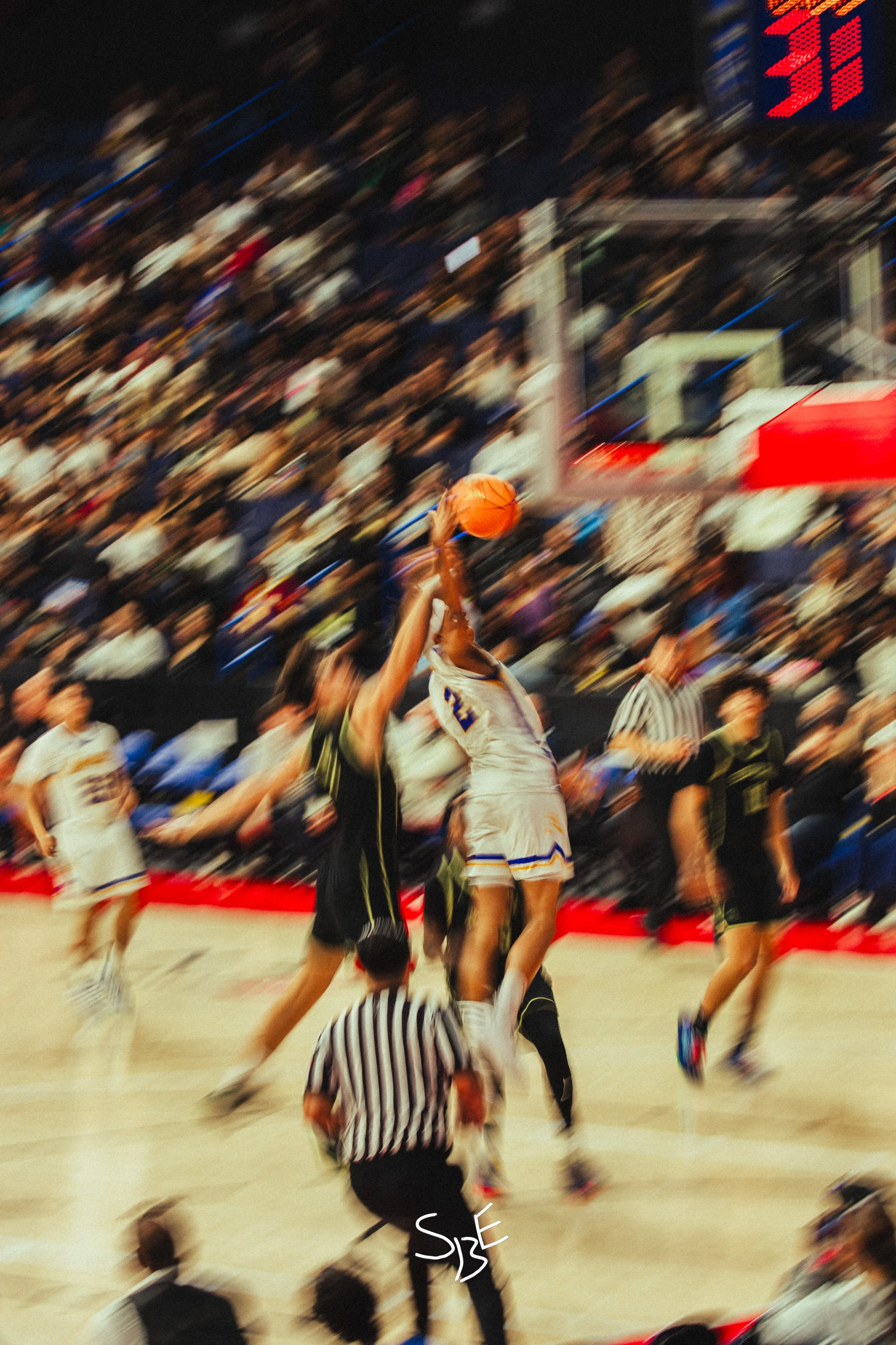 A referee, in a black and white striped shirt, is on the basketball court at a basketball game. Two players are jumping for the ball, with one player wearing a white jersey and another in black defending. The crowd is in the stands, and the game is a