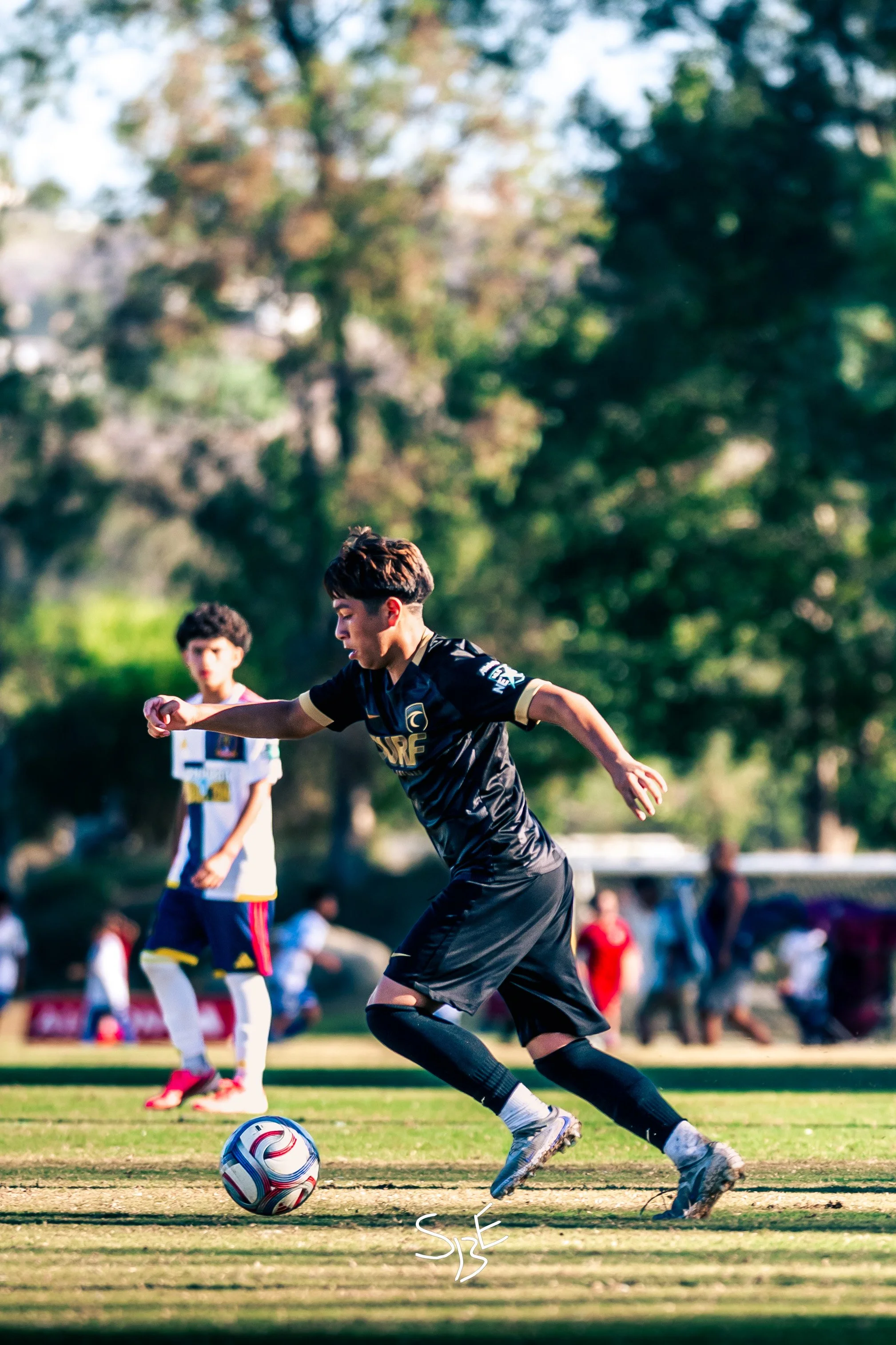 A soccer player in black uniform with the word 'FURE' on it is kicking a soccer ball on a field. In the background, there is a person in a white jersey and several other people, with trees and sky visible.