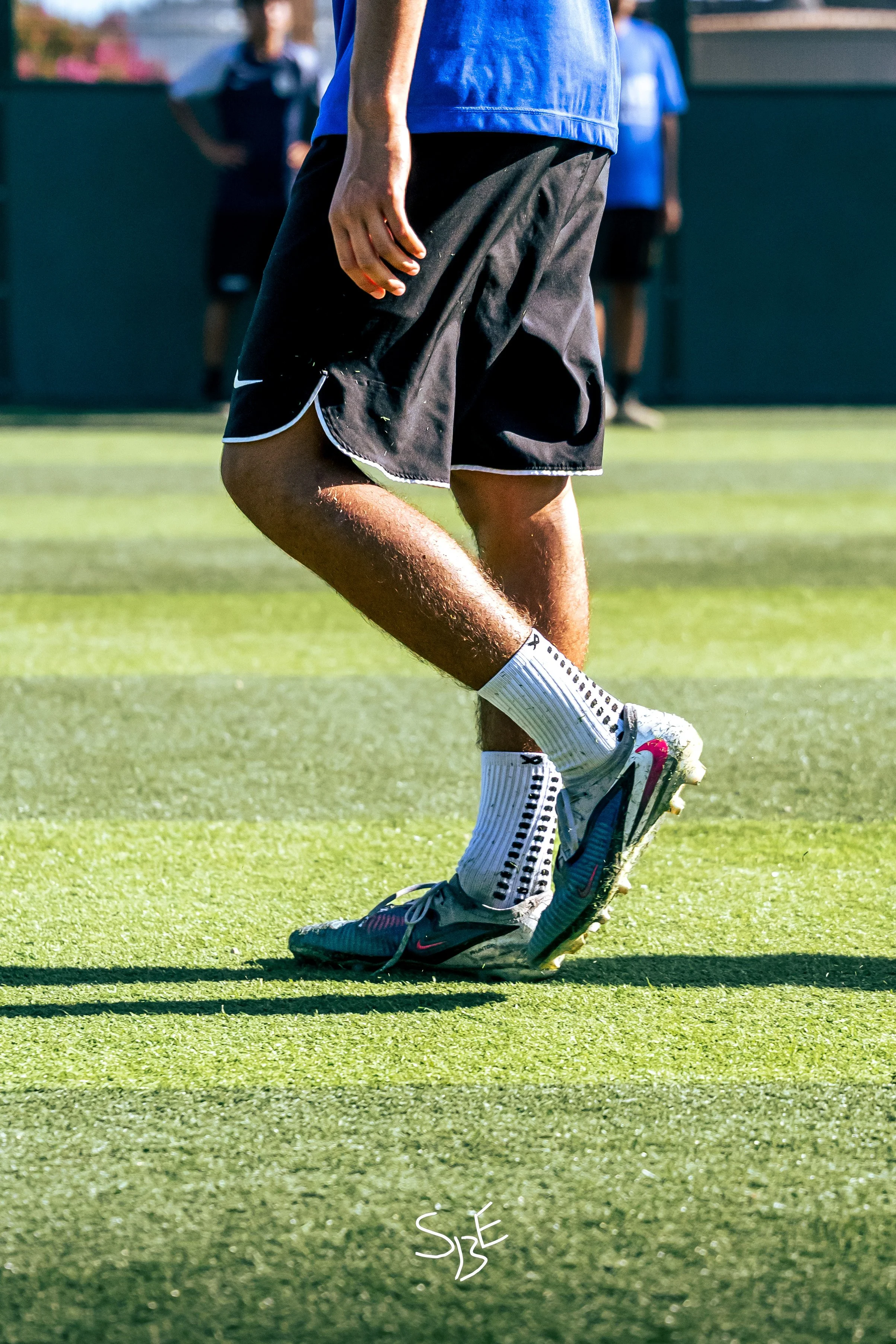 Close-up of a person standing on a sports field, wearing athletic shoes and black shorts, with a blurred background of other people and a green fence.