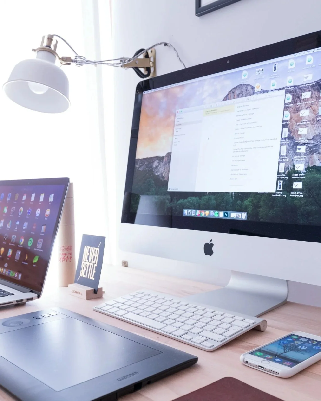 A modern workspace with an Apple iMac desktop computer, a laptop, a wireless keyboard, a wireless mouse, a smartphone, and various desk accessories. A sleek, minimalist style with white and wooden elements.