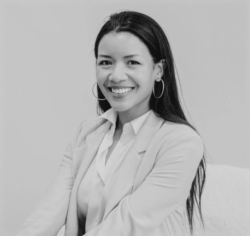 A portrait of a woman with long dark hair, wearing a light-colored blazer and hoop earrings, smiling, in black and white.