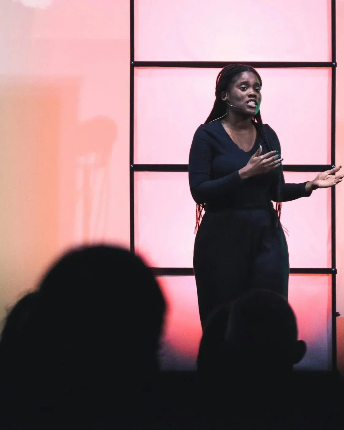 A woman giving a presentation in front of a pink backdrop with black grid framing, speaking to an audience.