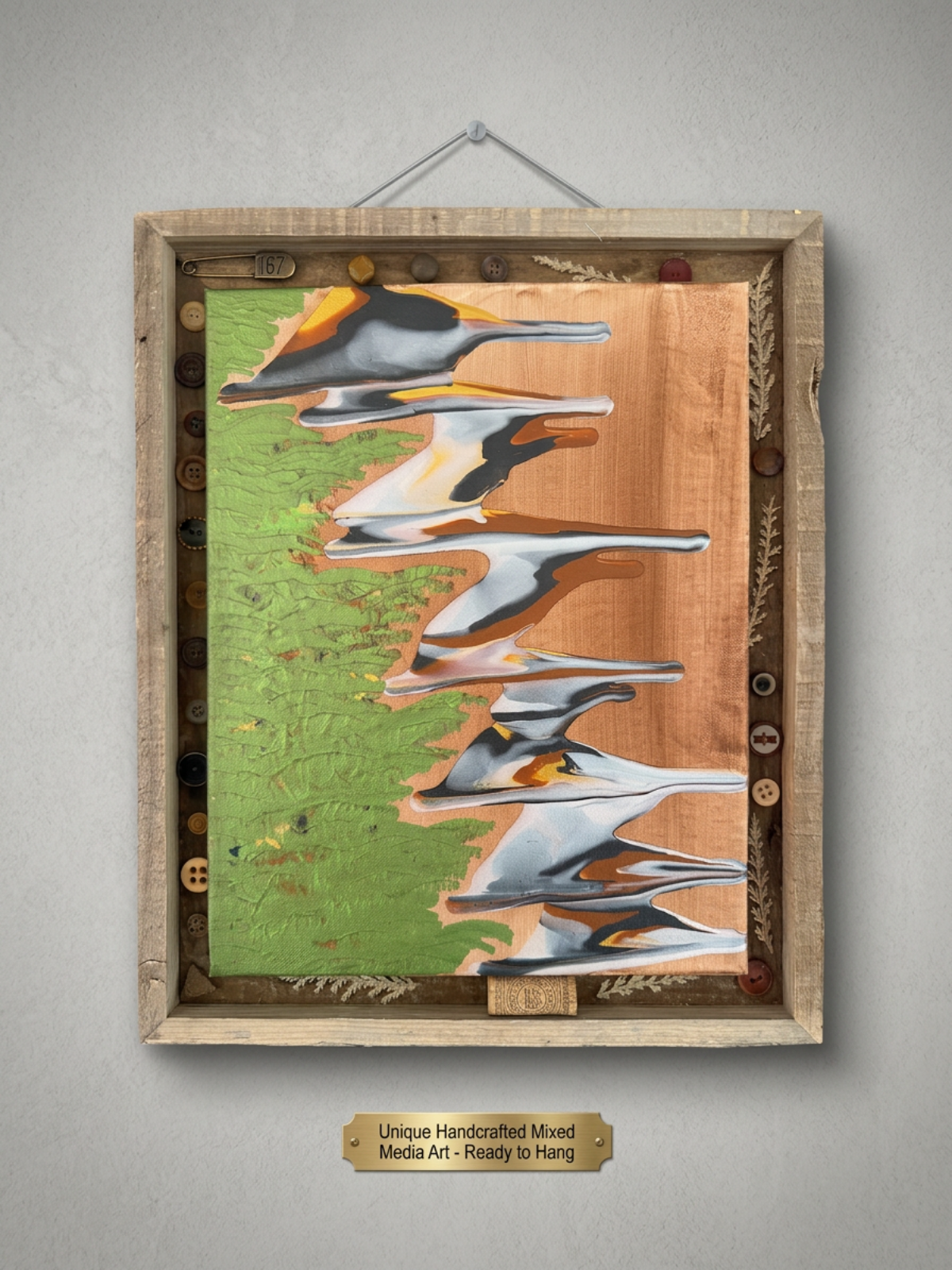 Handcrafted mixed media art featuring a painting of a mountain landscape with a river and green trees, framed by a wooden box decorated with various buttons and a small label at the bottom reading "Unique Handcrafted Mixed Media Art - Ready to Hang."