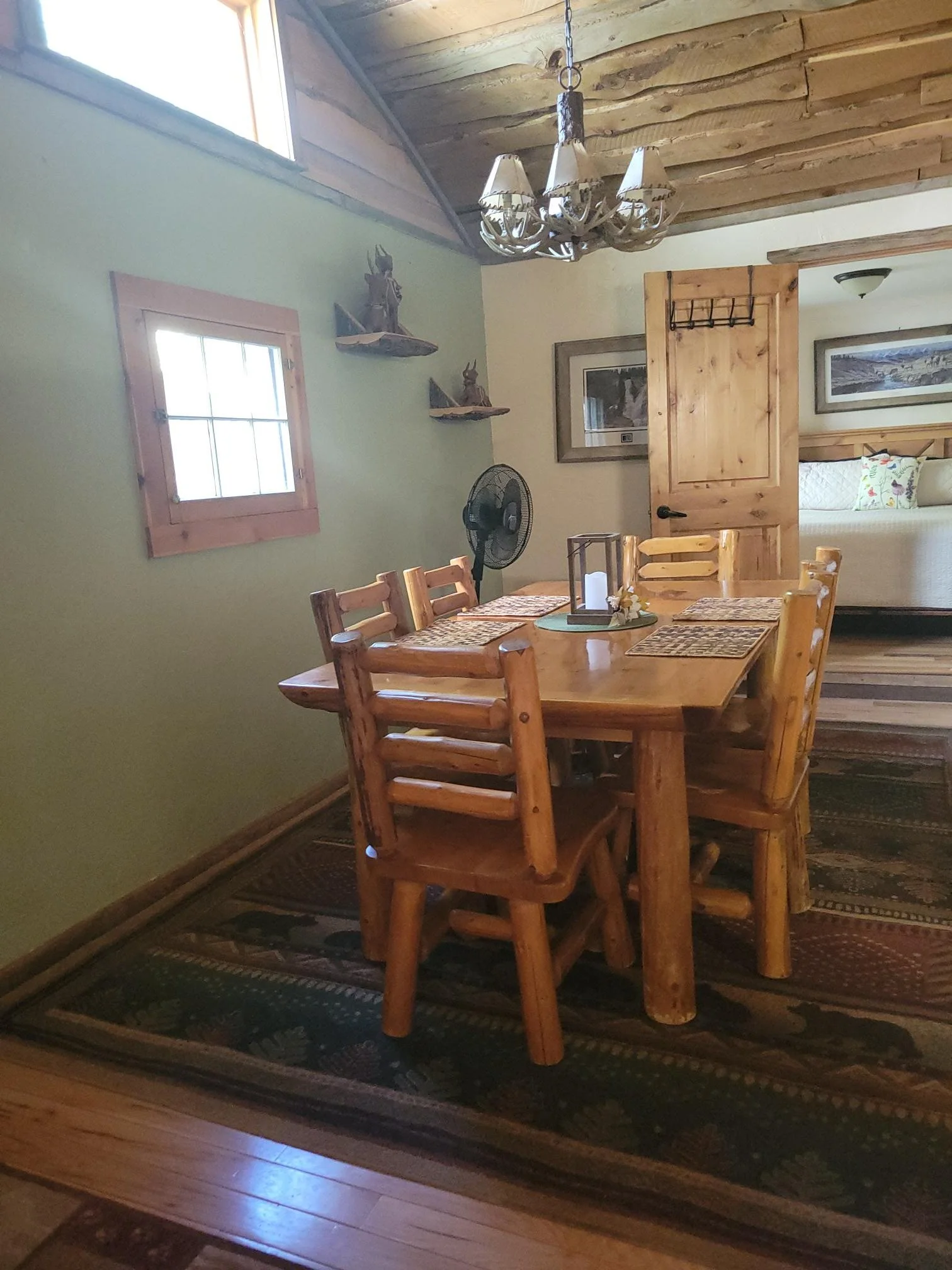 Interior of a rustic dining room with a wooden table, matching chairs, a chandelier made of antlers, framed pictures on the wall, a fan, and a partially open wooden door leading to a bedroom with a bed and decorative pillows.
