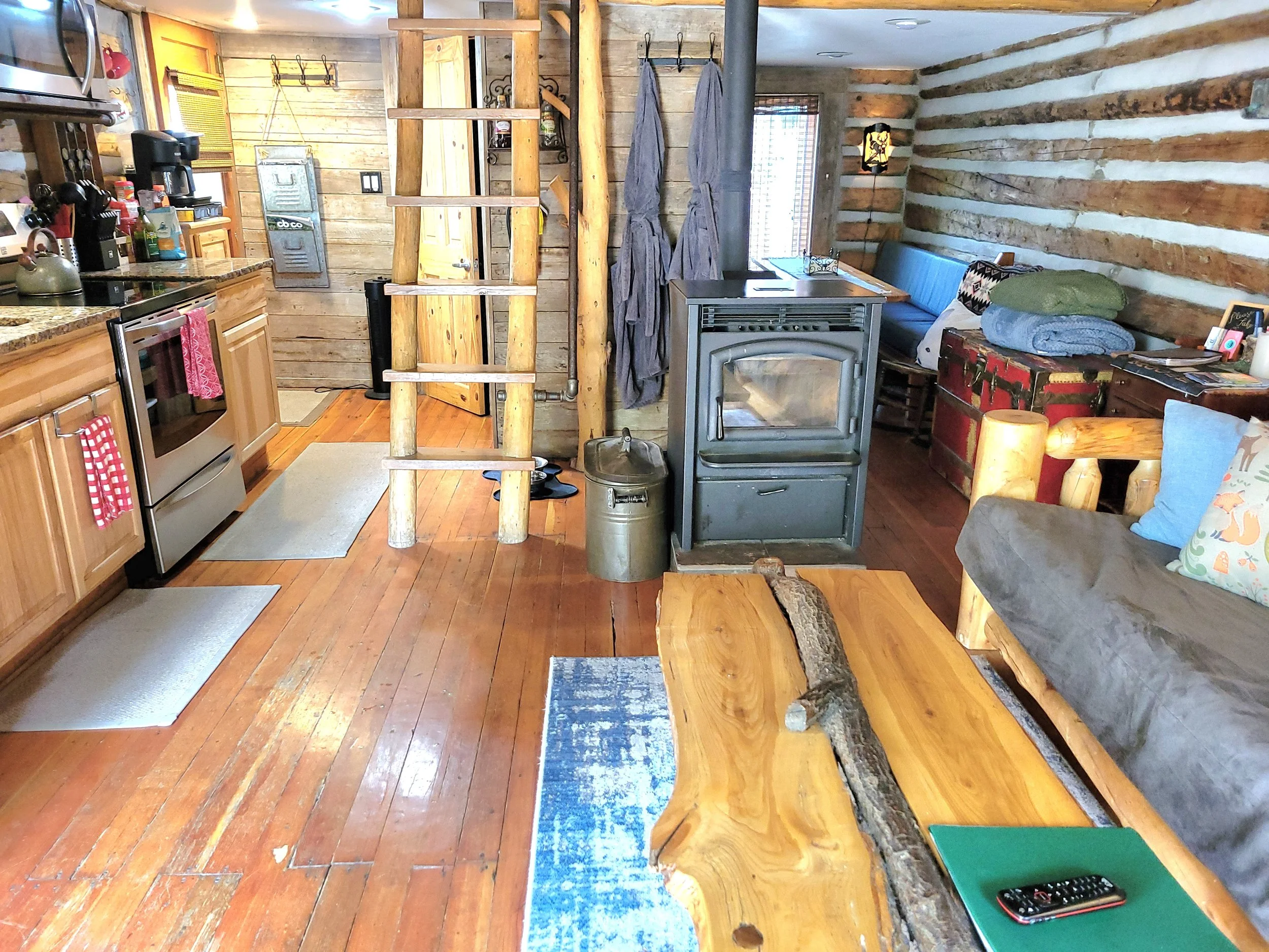 Interior of a rustic cabin with wooden walls and floor, featuring a kitchen area with appliances and a living space with a bench, a wood stove, and a log table.