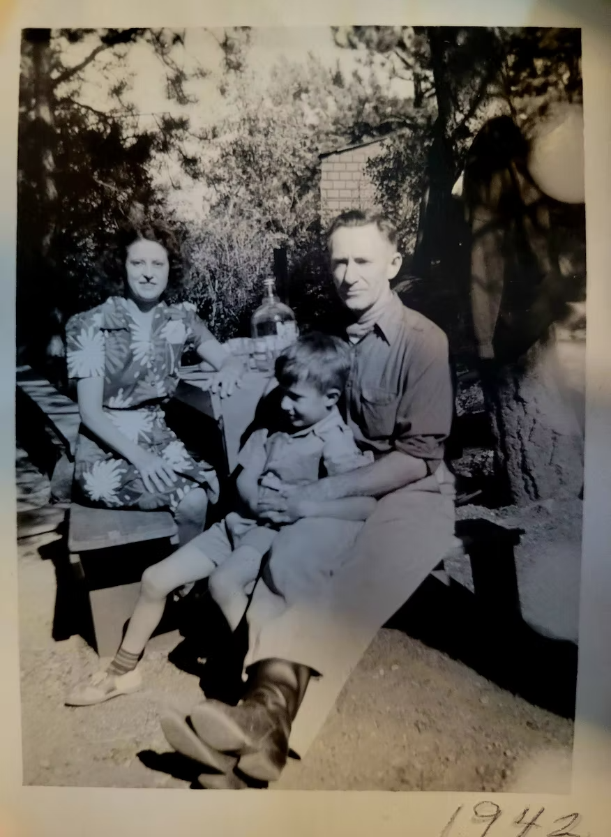 A black and white photo of a family photo outdoors. A woman and man sit with a young boy on their laps. The woman wears a floral dress, and the man wears a buttoned shirt. The woman sits on a wooden bench next to a woman, and there is a bottle on the table behind them. Trees, a brick structure, and a person with a balloon are in the background. The year 1942 is handwritten in the bottom right corner.