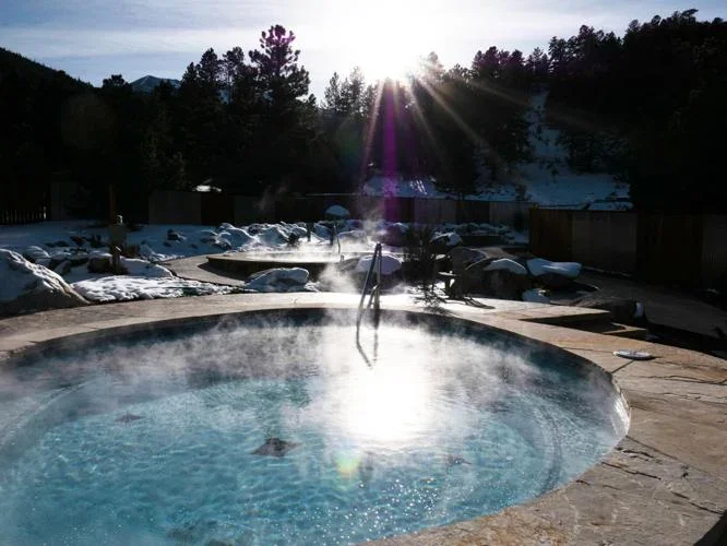 Outdoor hot tub with bubbling water, surrounded by snow on the ground, with the sun shining over trees in the background.