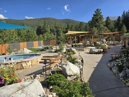 Scenic outdoor area with a pool, colorful shade sails, a wooden picnic shelter, stone landscaping, and lush green trees with mountains in the background.