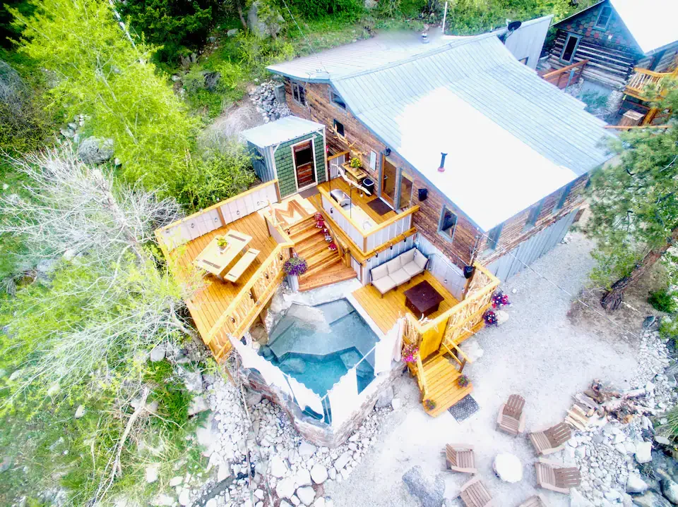 Aerial view of a rustic cabin with a deck, small pool, outdoor seating, surrounded by trees and rocks.