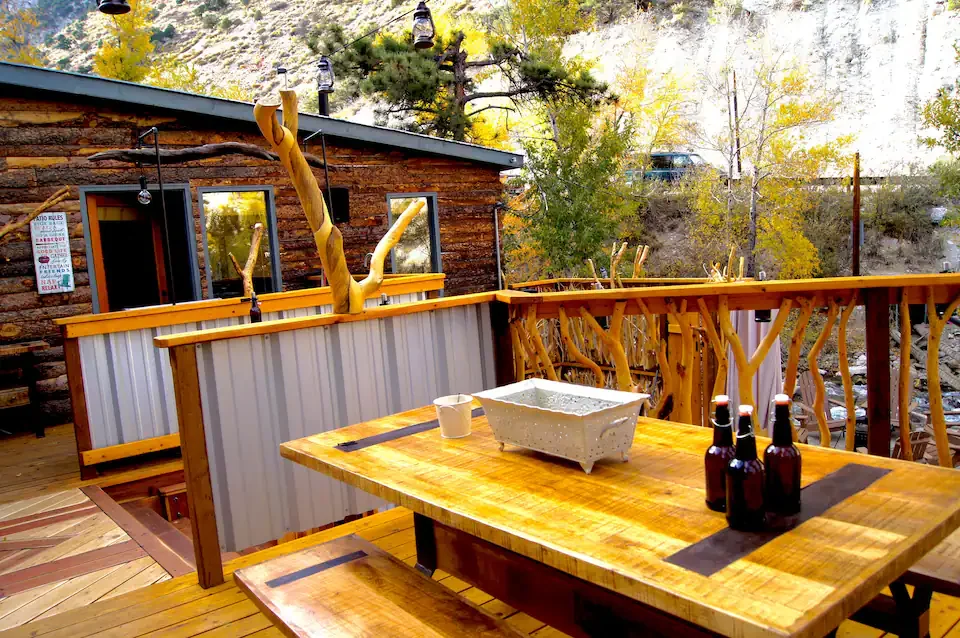 Wooden deck with a rustic railing, a table with three bottles, a mug, and a planter, outdoor setting with trees and a building in the background.
