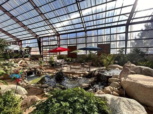 Indoor tropical garden with rocks, a small waterfall, plants, and seating area with tables and umbrellas.