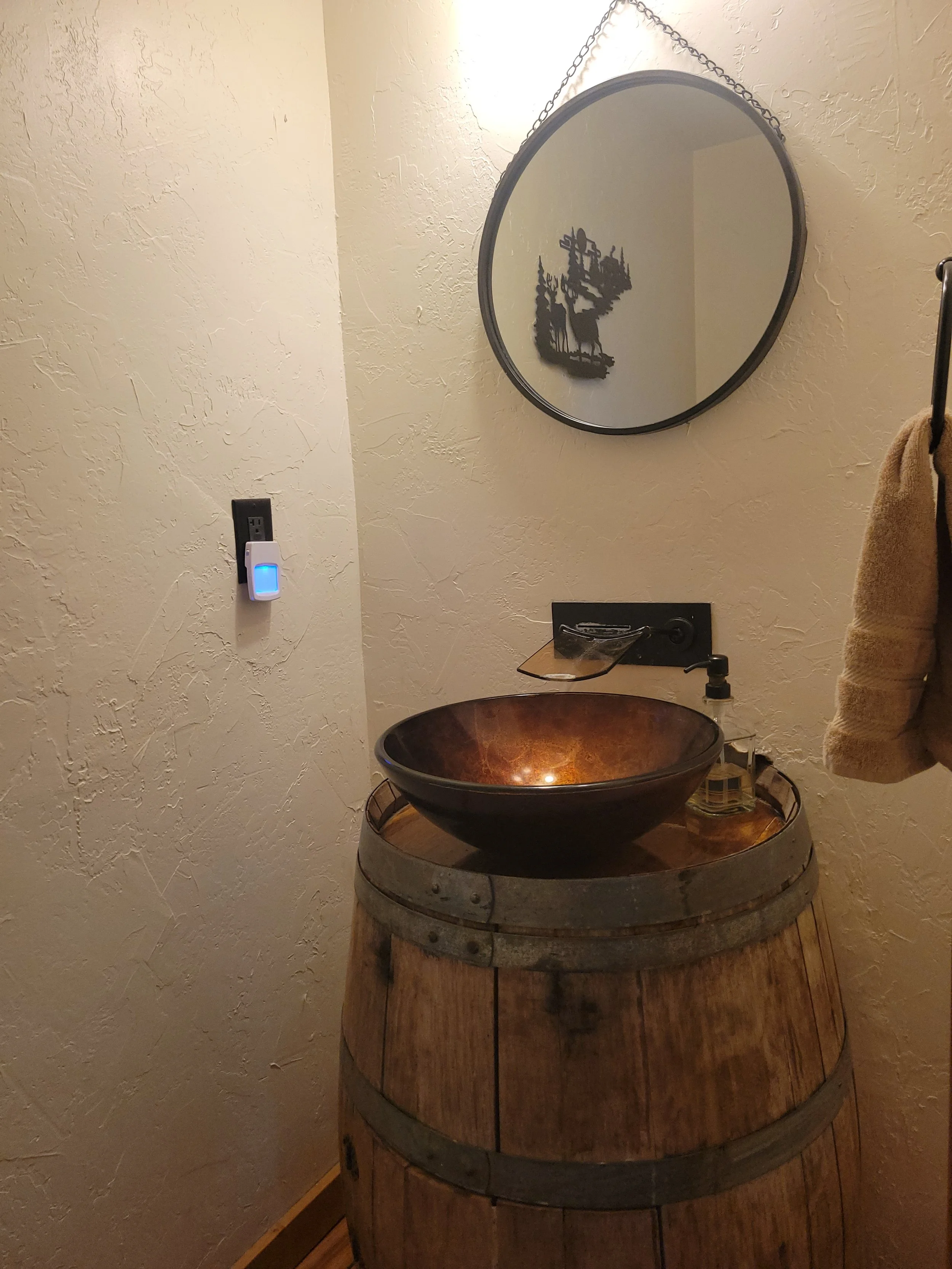 A rustic bathroom vanity made from a large wooden barrel with a copper bowl sink on top, a bottle of soap, and a black faucet. There is a round mirror with a chain hanging on the textured beige wall above the vanity. A towel hangs to the right side of the image.