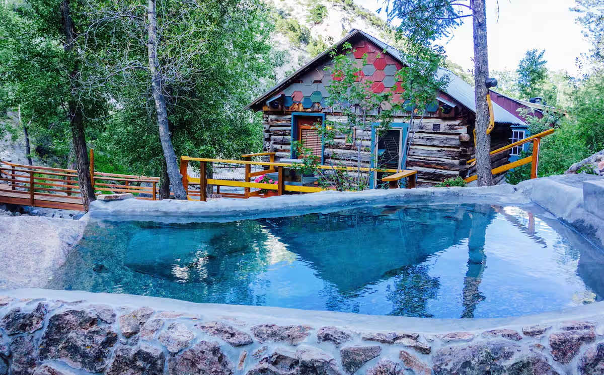 Log cabin with a sloped roof, surrounded by trees, with a small balcony and flowers, adjacent to an outdoor swimming pool with rocky edges, in a forested area.
