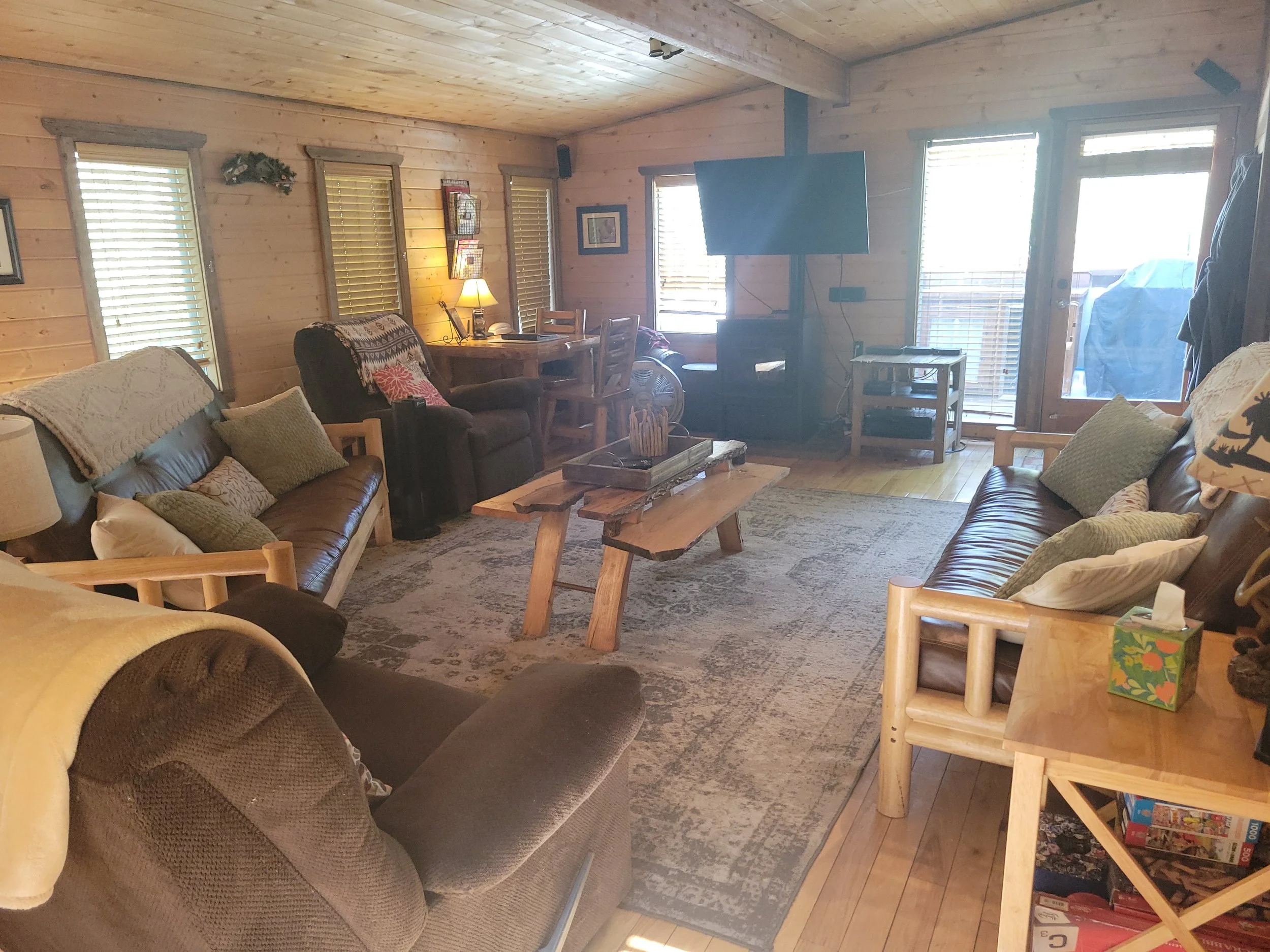Living room with wooden walls and ceiling, featuring multiple windows with blinds, a television mounted on the wall, a sitting area with sofas and pillows, a wooden coffee table, a dining table with chairs, and a door leading outside. There are lamps, pictures, and various decorative items around the room.
