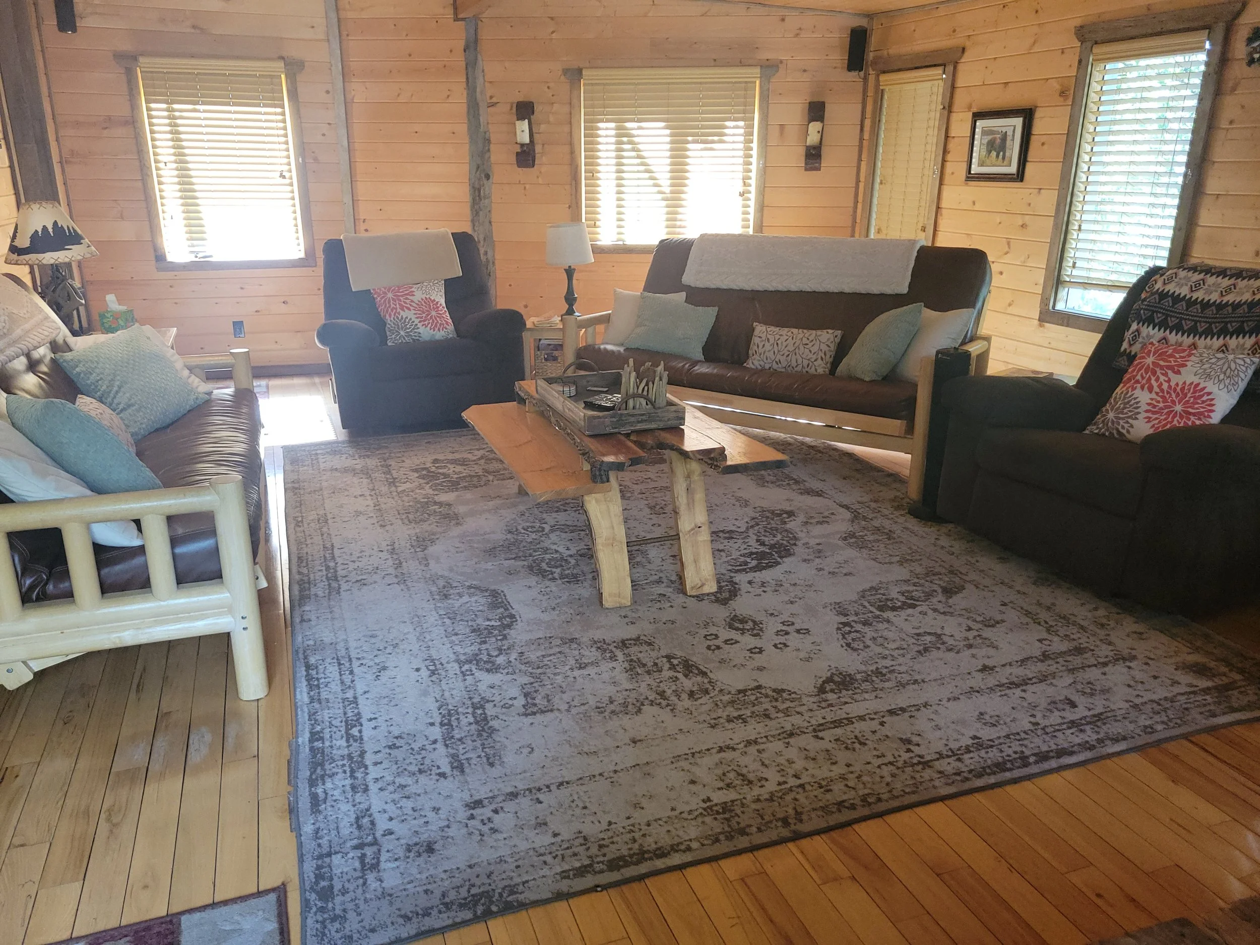 A cozy living room with wooden walls and multiple windows covered by blinds. It features a large area rug, a coffee table, and several chairs and sofas adorned with pillows and throws.