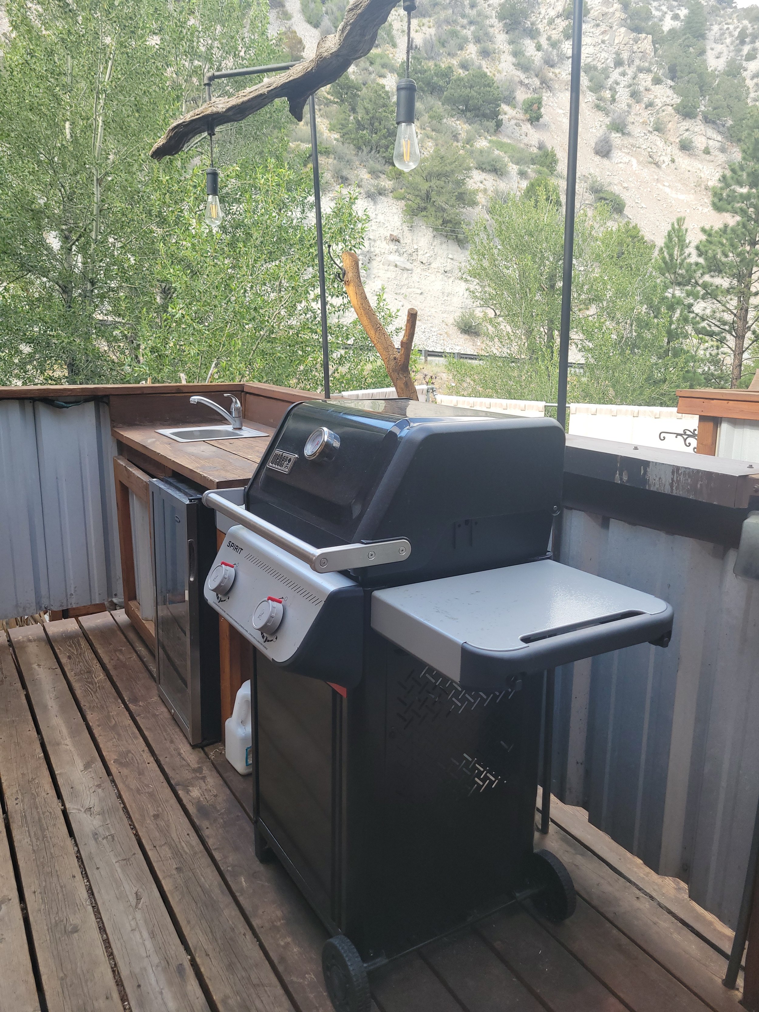 An outdoor patio area with a black gas grill, a wooden counter with a small sink, and a string of light bulbs hanging from a tree branch. The background shows a hillside with trees and rocky terrain.