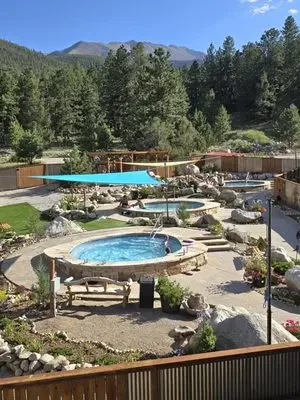Outdoor pool area with two hot tubs, a small pool, and a poolside shade sail, surrounded by trees and mountainous landscape in the background on a sunny day.