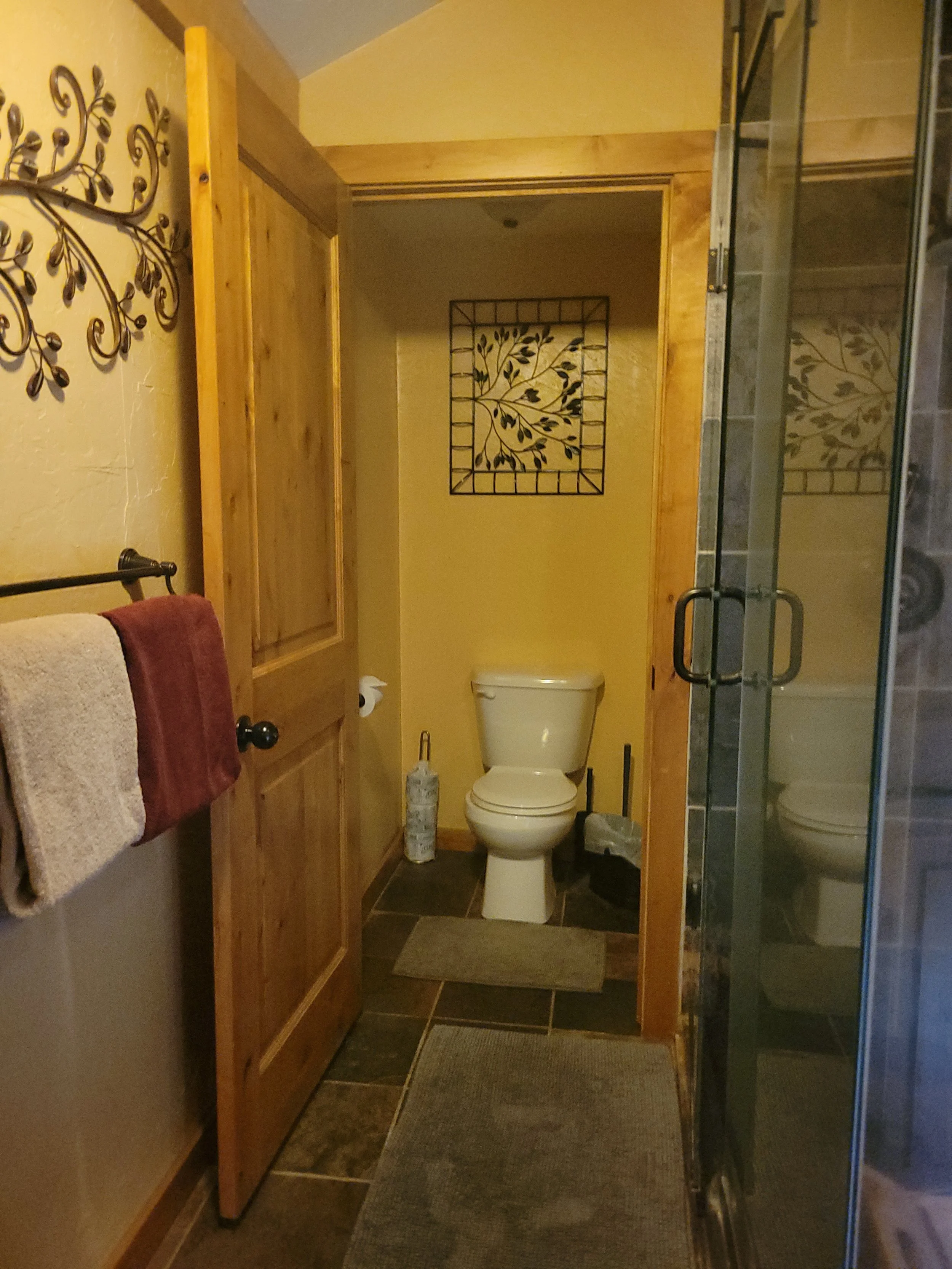 A small bathroom with a toilet positioned against the back wall, with a narrow wooden door to the left and a glass shower stall to the right. The back wall has a decorative metal wall piece with a leaf pattern. There are towels hanging on a black metal towel rack on the left wall.