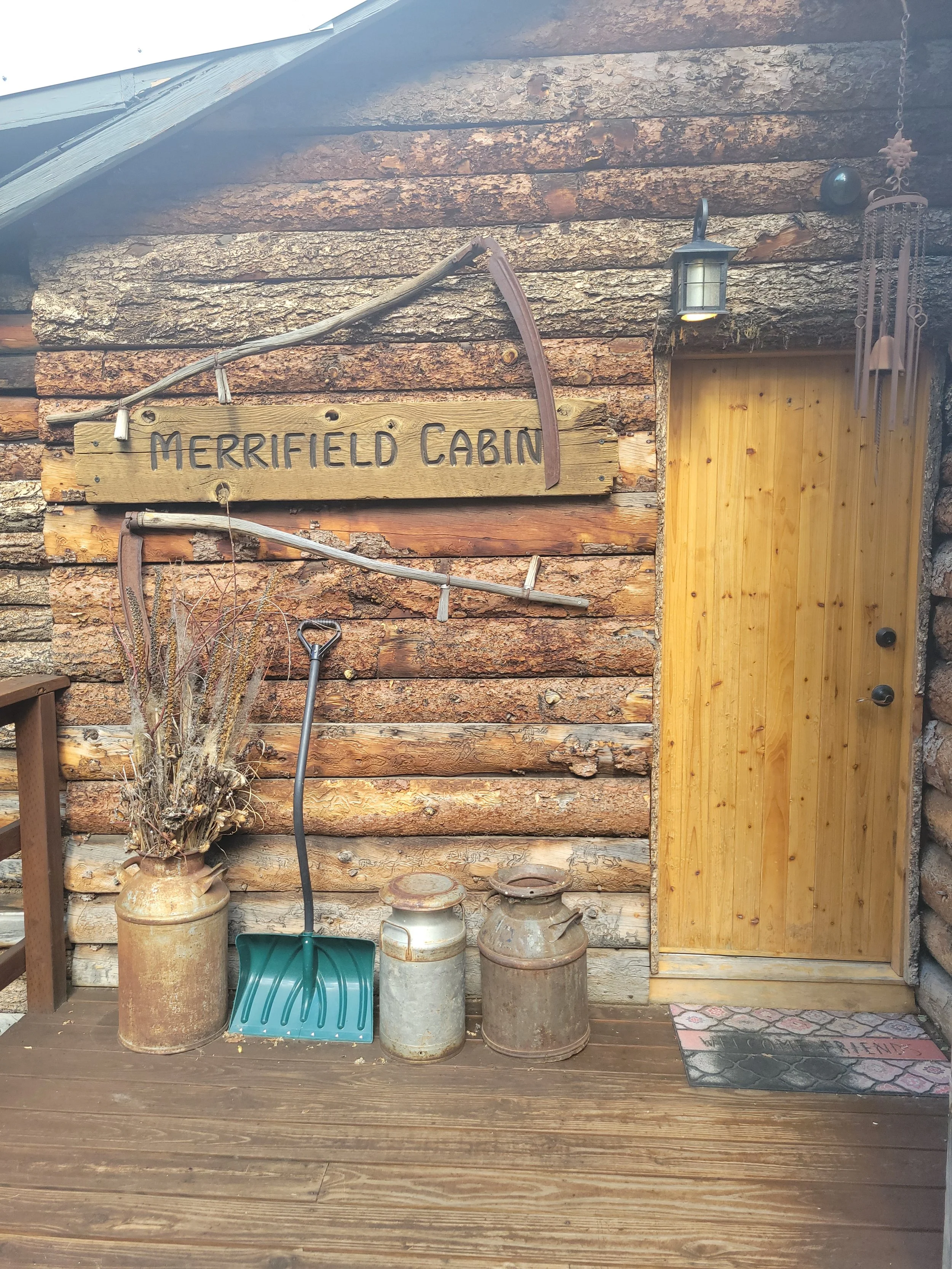 Wooden cabin exterior with a sign that reads 'Merrified Cabin', outdoor lights, wind chimes, and a door with a welcome mat. In front, there are old milk cans, a small green shovel, and a potted plant.