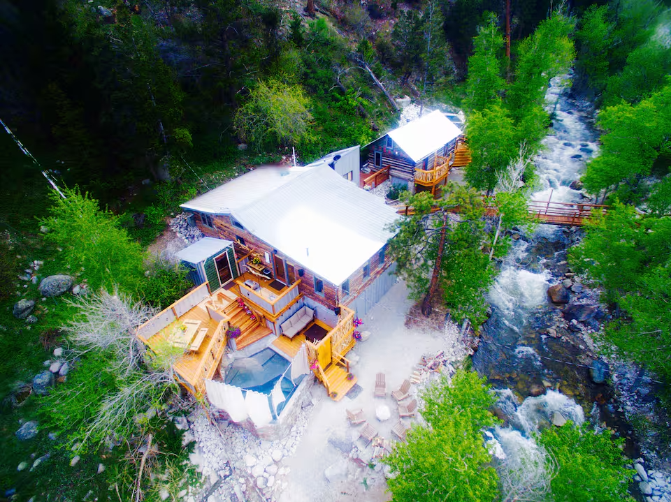 Aerial view of a cabin with multiple decks by a river in a forested area.