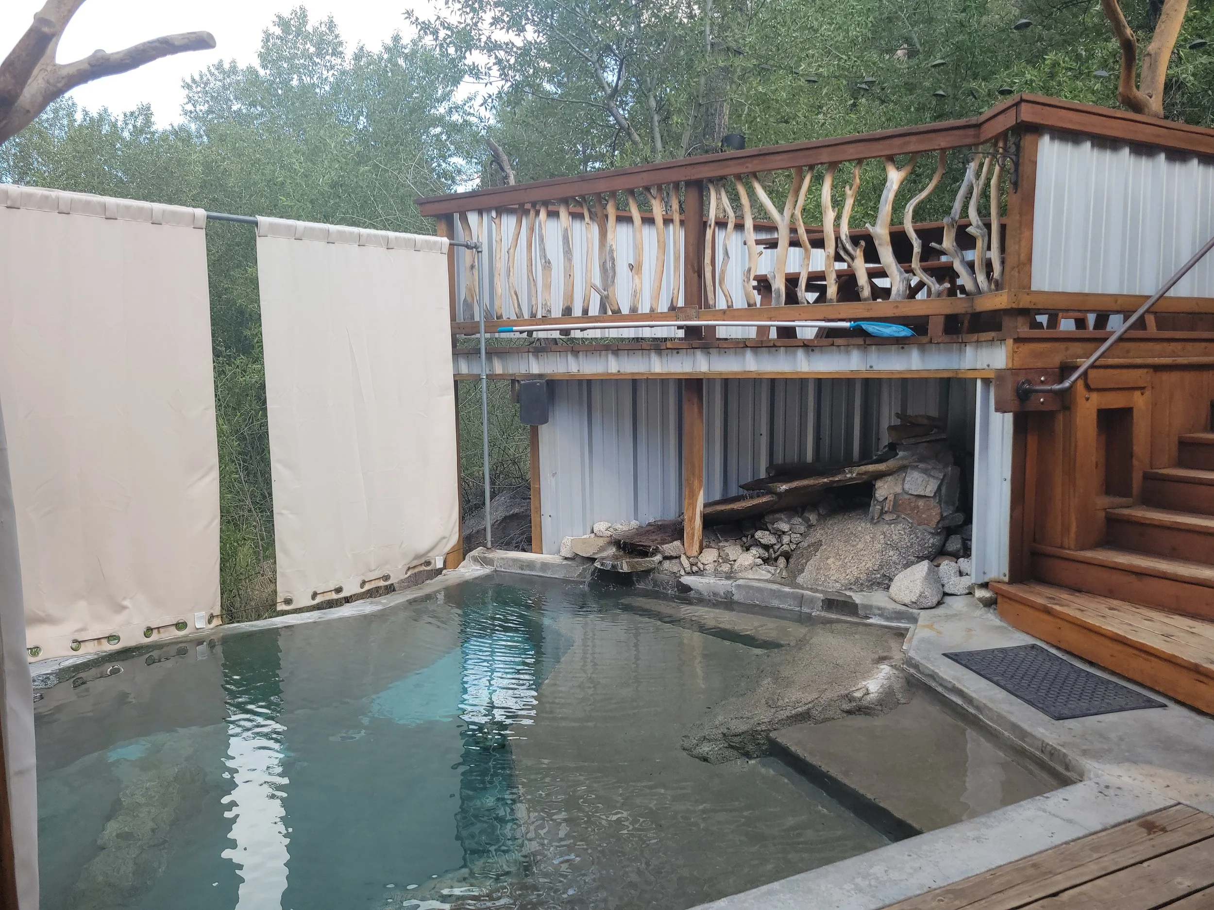 Outdoor hot spring with a concrete edge, surrounded by trees. A stone and wood structure, possibly a small waterfall or water feature, is on the right, with a staircase leading up. A white fabric privacy curtain is on the left, and the background shows lush green trees.