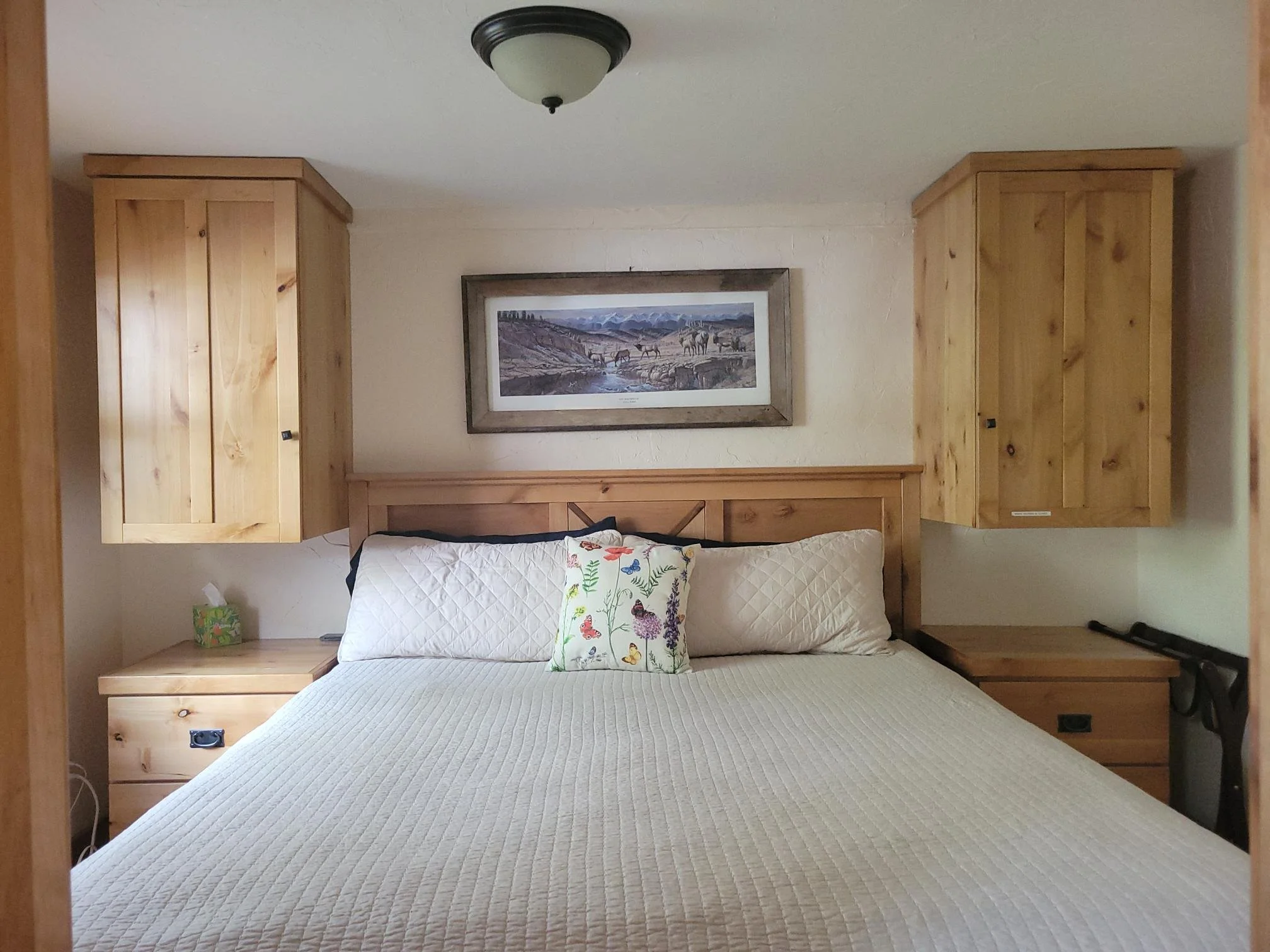 A cozy bedroom with a bed covered by a beige quilt and a decorative pillow with floral and butterfly patterns. The headboard is wooden, and there's a bedside table with a small green and orange globe lamp on the left. Wooden cabinets are mounted on the wall behind the bed, and a framed landscape painting hangs above the headboard. A ceiling light fixture is also visible.