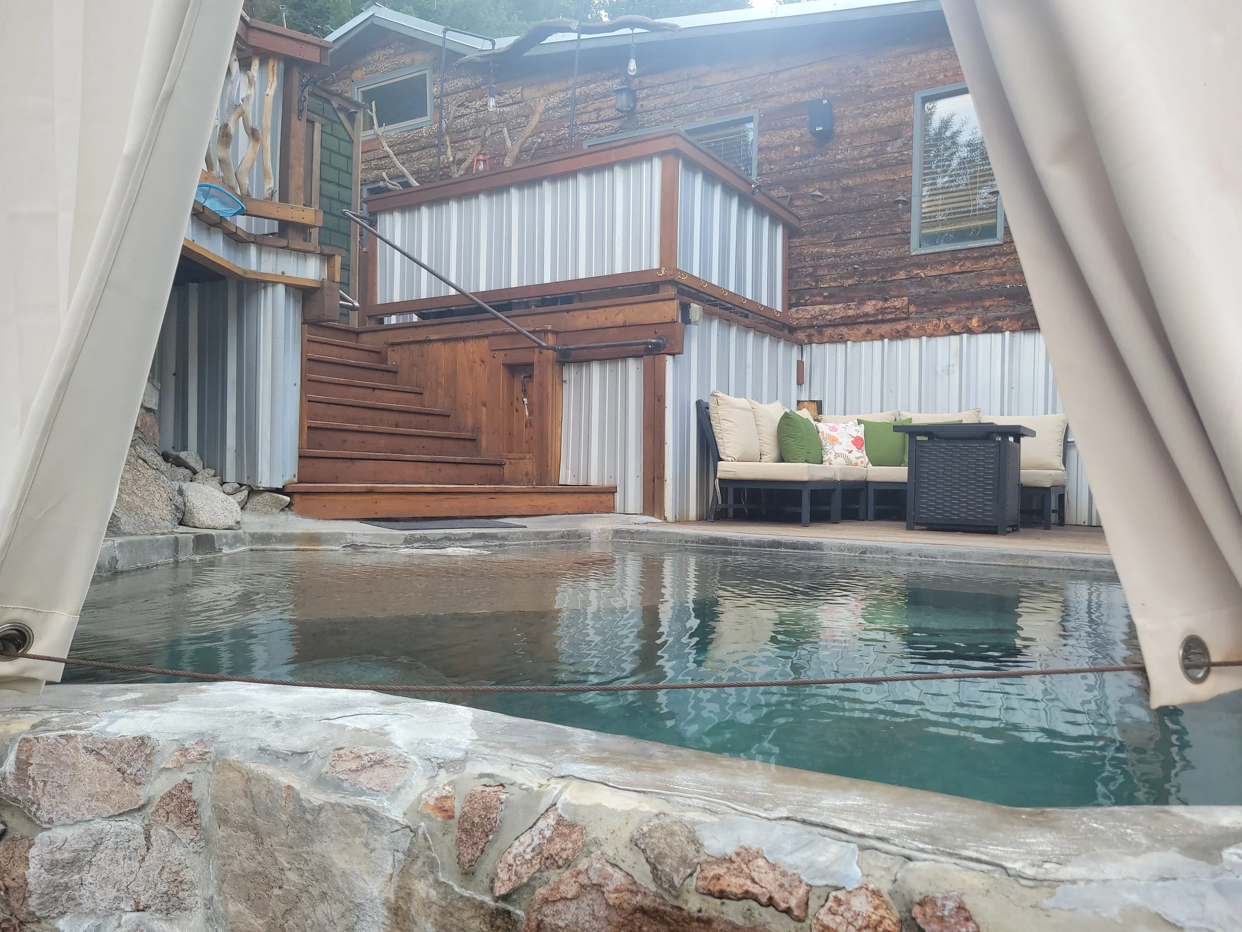 View from inside a hot tub looking out onto a backyard patio with outdoor furniture, wooden stairs, a wooden and metal structure, and a wooden house with windows and string lights.