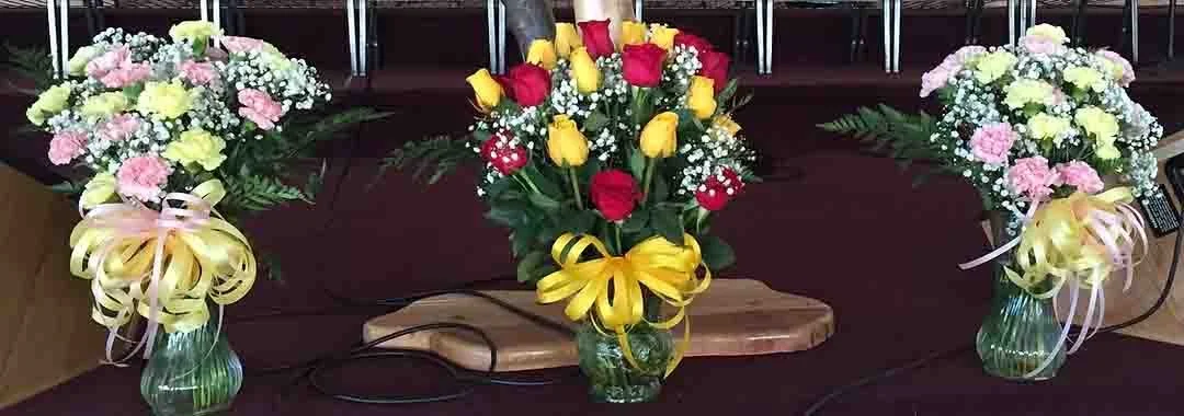 Three bouquets of flowers in vases, with the middle bouquet featuring yellow and red roses, and the other two bouquets with pink and white flowers, on a dark surface.