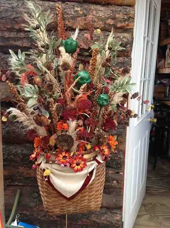 Decorative floral arrangement in a woven basket with autumn-colored flowers, dried leaves, and artificial green pumpkins, set against a stone wall and partially open white door.