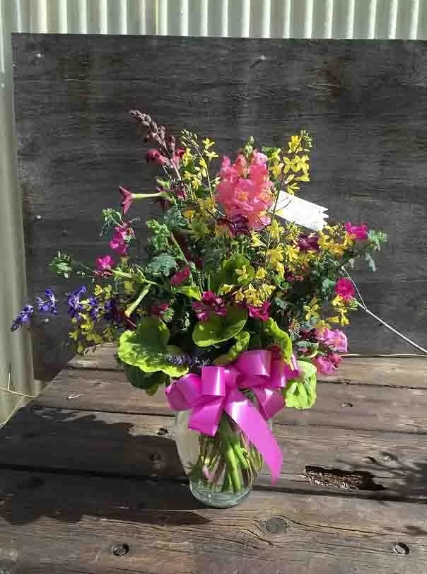 A glass vase with a colorful assortment of flowers, tied with a pink ribbon, placed on a wooden surface.
