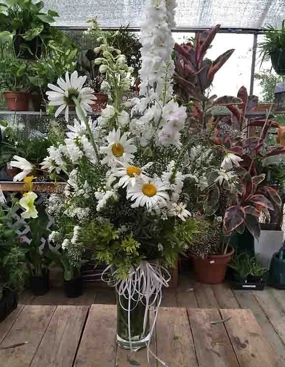 A large bouquet of white daisies and other white flowers in a tall glass vase set on a wooden surface, with potted plants and greenery in the background.