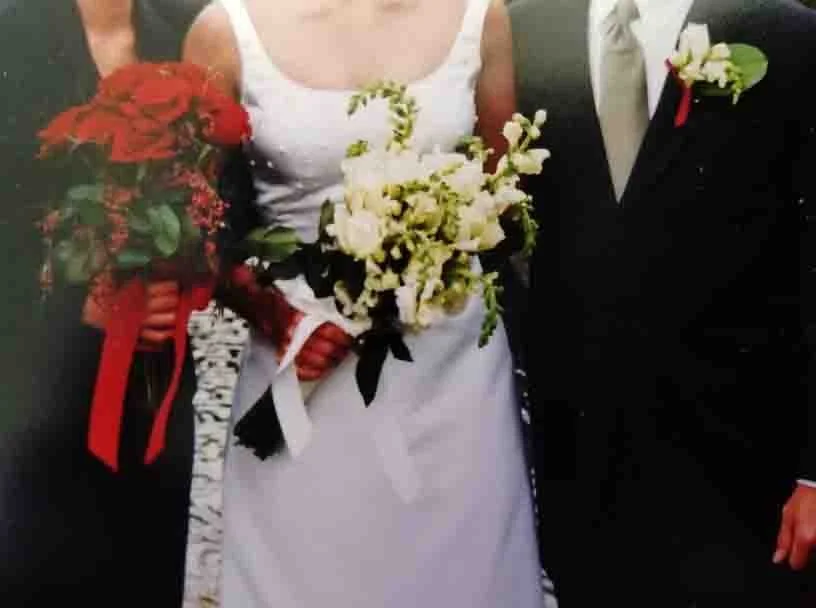 Bride holding a bouquet of white flowers, wearing a white wedding dress. Groom wearing a black suit and white dress shirt, with a white boutonniere and a red ribbon.