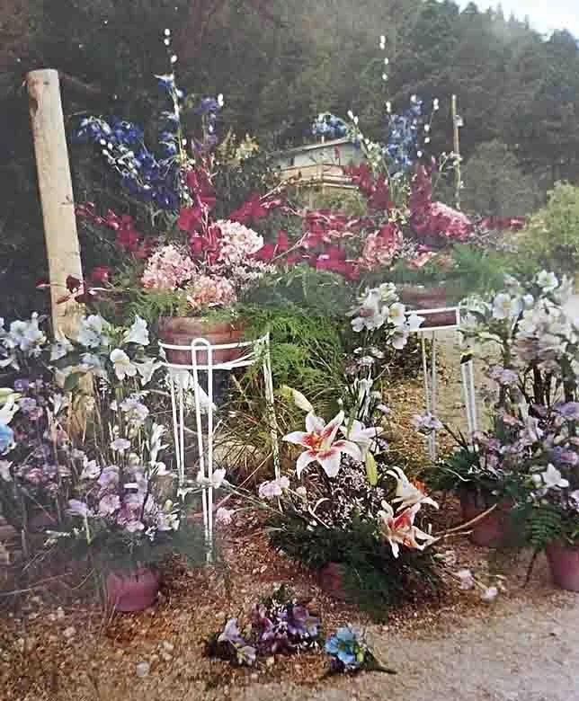 Flower arrangement outdoors with pink, purple, and white flowers, including lilies, set on white stands and surrounding a gravel path, with trees and a building in the background.
