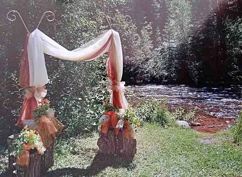 Outdoor wedding ceremony setup with a fabric arch decorated with flowers, near a river in a wooded area.