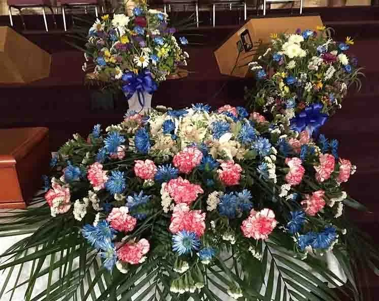 Large floral arrangement with pink, blue, white, and yellow flowers, accompanied by broad green leaves, in a church or auditorium setting.