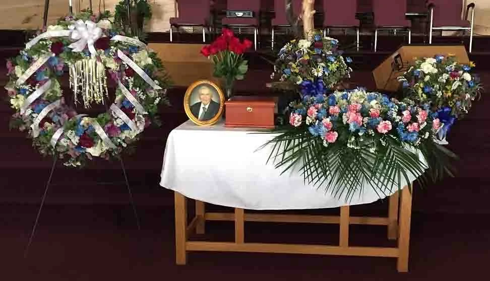 Funeral arrangement with colorful flowers, a framed photograph of a man, and a wooden box on a white-draped table.