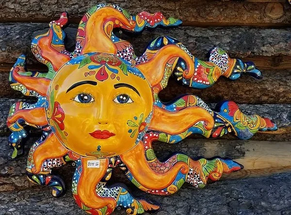 Colorful ceramic wall decoration of a yellow sun with a human face, surrounded by multicolored, swirling rays, mounted on a wooden wall.