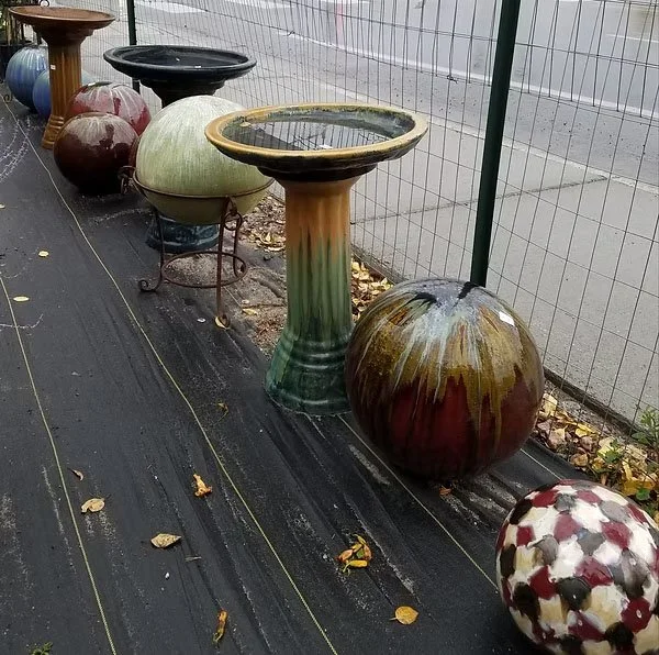 Various ceramic garden pots, some with water, arranged on a black tarp outdoors, behind a wire fence.