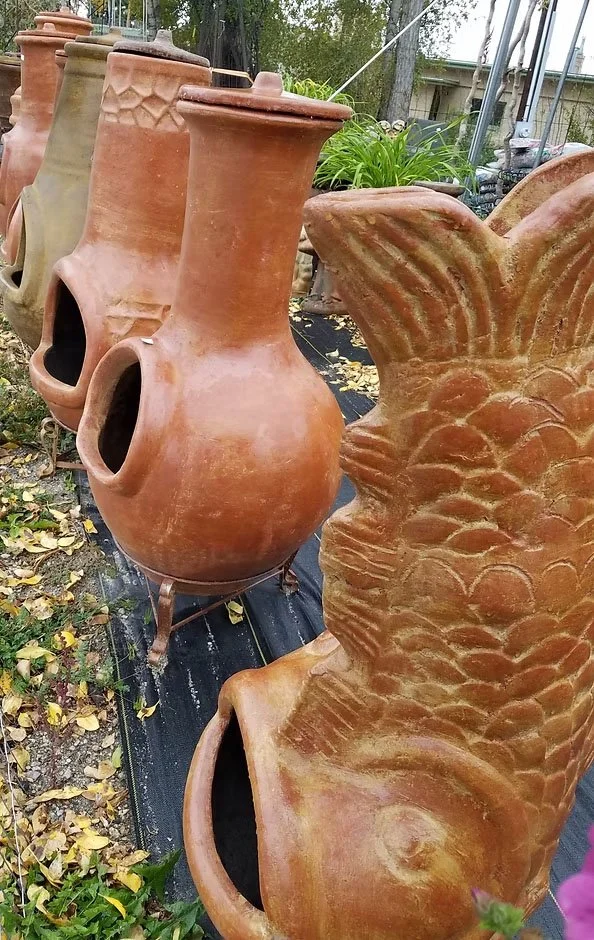 Several large terracotta garden planters with openings at the front, some standing on metal stands, outdoors with green plants and trees in the background.