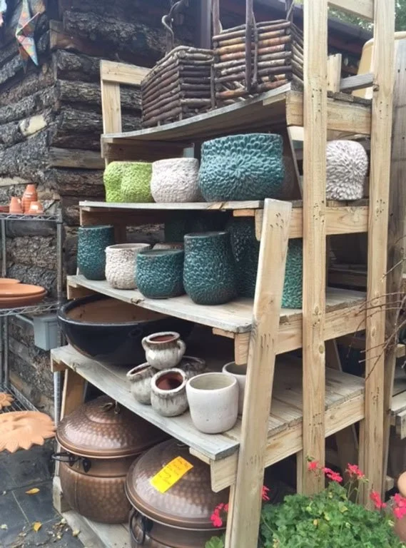 Wooden outdoor shelving unit displaying various ceramic and clay pots, including textured planters, bowls, and large urns, with some gardening tools and decorations visible around.