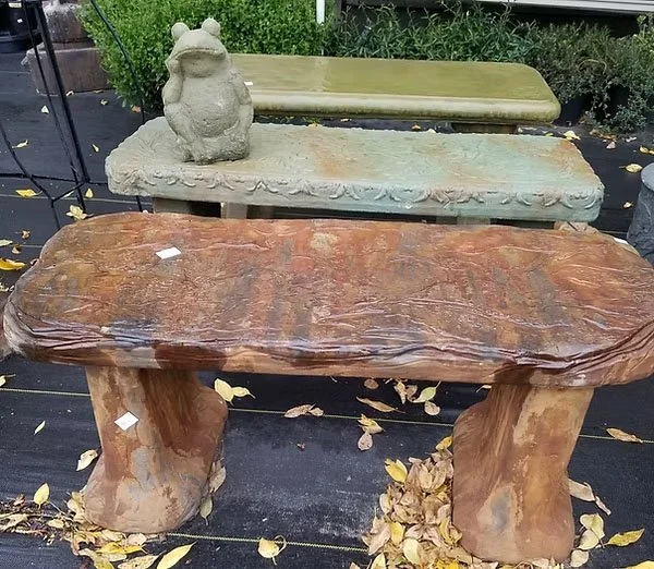 Two carved stone benches outdoors, one in front with a polished brown finish and one in the background with a rough gray finish, surrounded by fallen leaves.