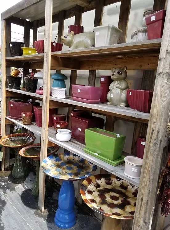 Shelves filled with various colorful ceramic and plastic planters and pots, with some decorative animal-shaped planters, along with a few small bowls, displayed outdoors on a wooden stand.