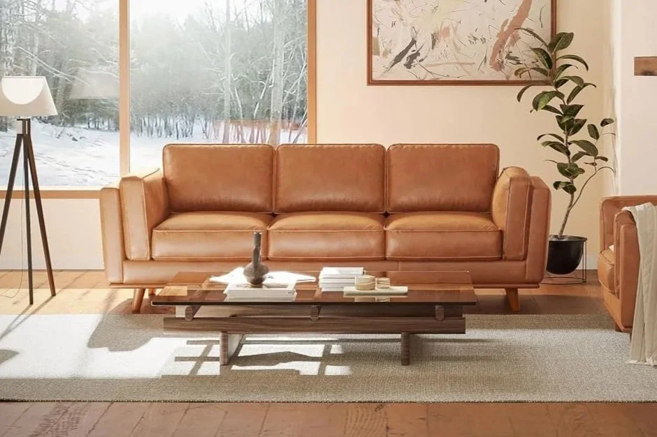 A living room with a tan leather mid-century sofa, a wooden coffee table, a large window showing a snowy landscape outside, a tall floor lamp with a white shade, an abstract painting on the wall, and a potted plant next to the sofa.