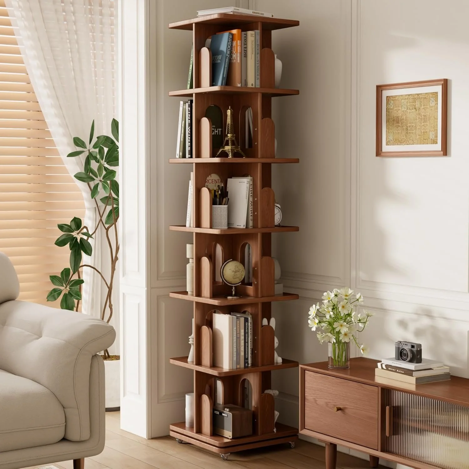 Living room corner with a wooden bookshelf holding books, decorative items, and a miniature Eiffel Tower. To the right, a wooden sideboard with a vase of white flowers, books, a camera, and a framed picture. To the left, part of a white sofa and a green leafy plant near a window with blinds and white curtains.