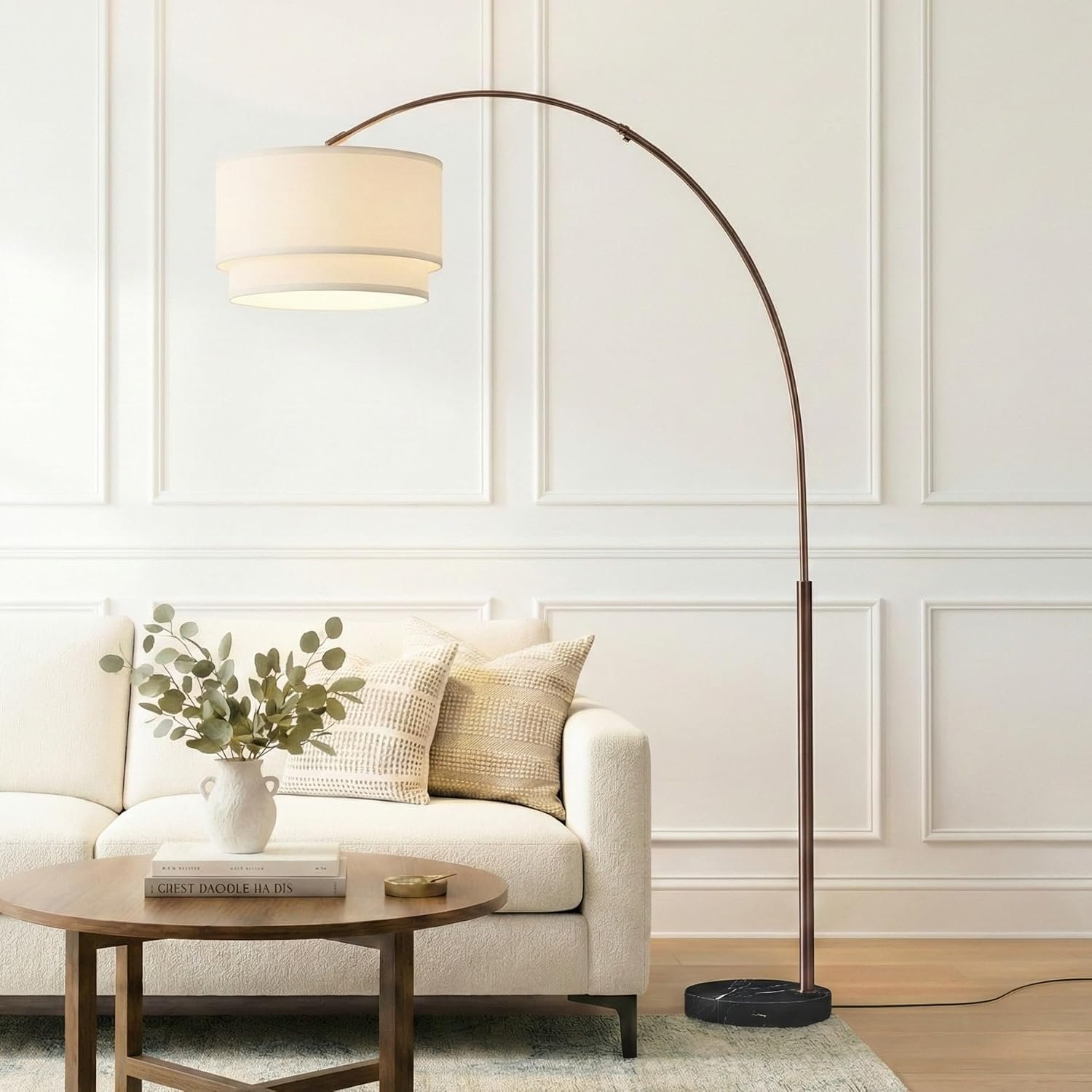 Living room with a cream sofa, beige cushions, a wooden coffee table with books and a vase of greenery, a white wall with decorative molding, and a modern floor lamp with a white shade and a black base.