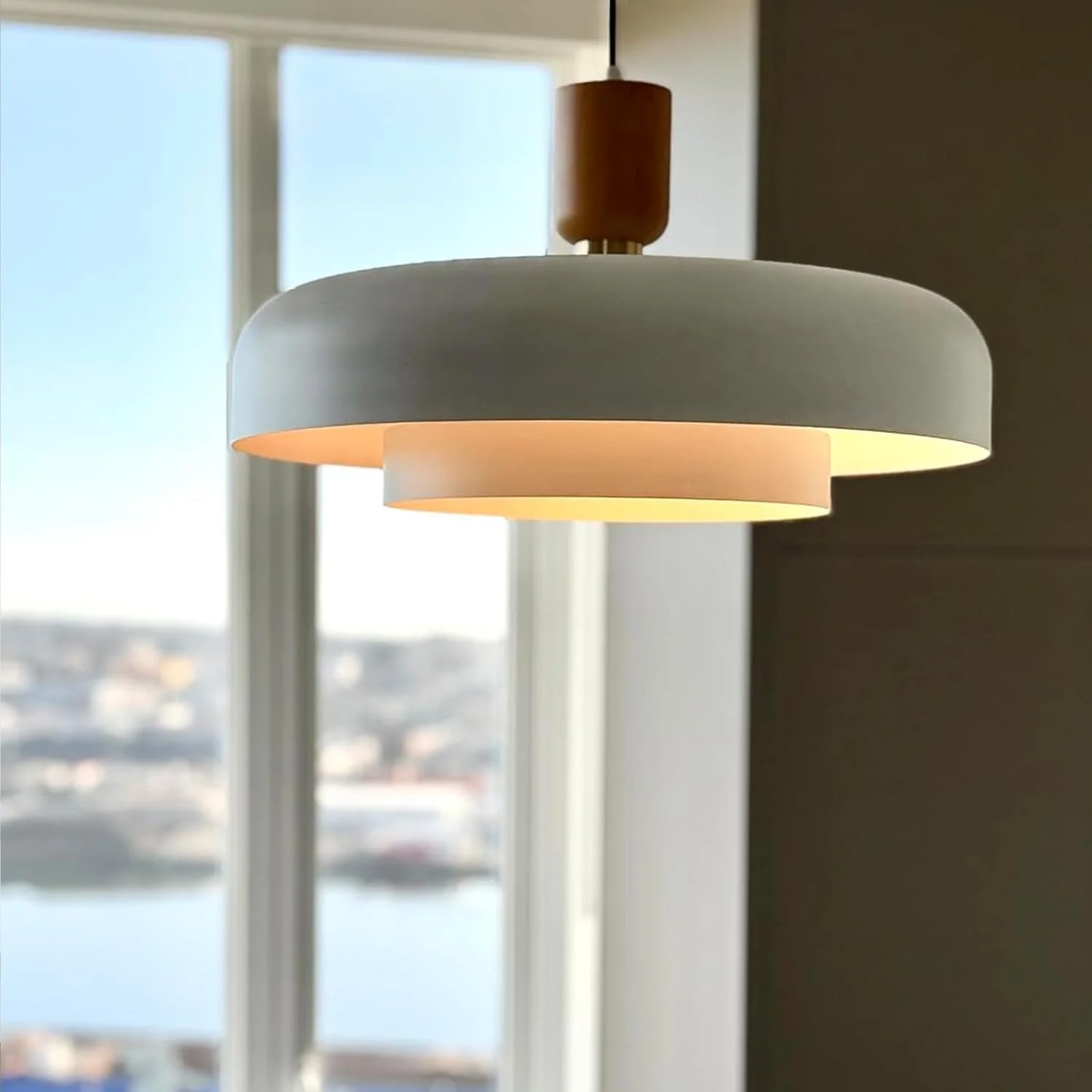 A modern hanging ceiling lamp with a white shade, wooden accent, and warm light in front of a window with a city view in the background.