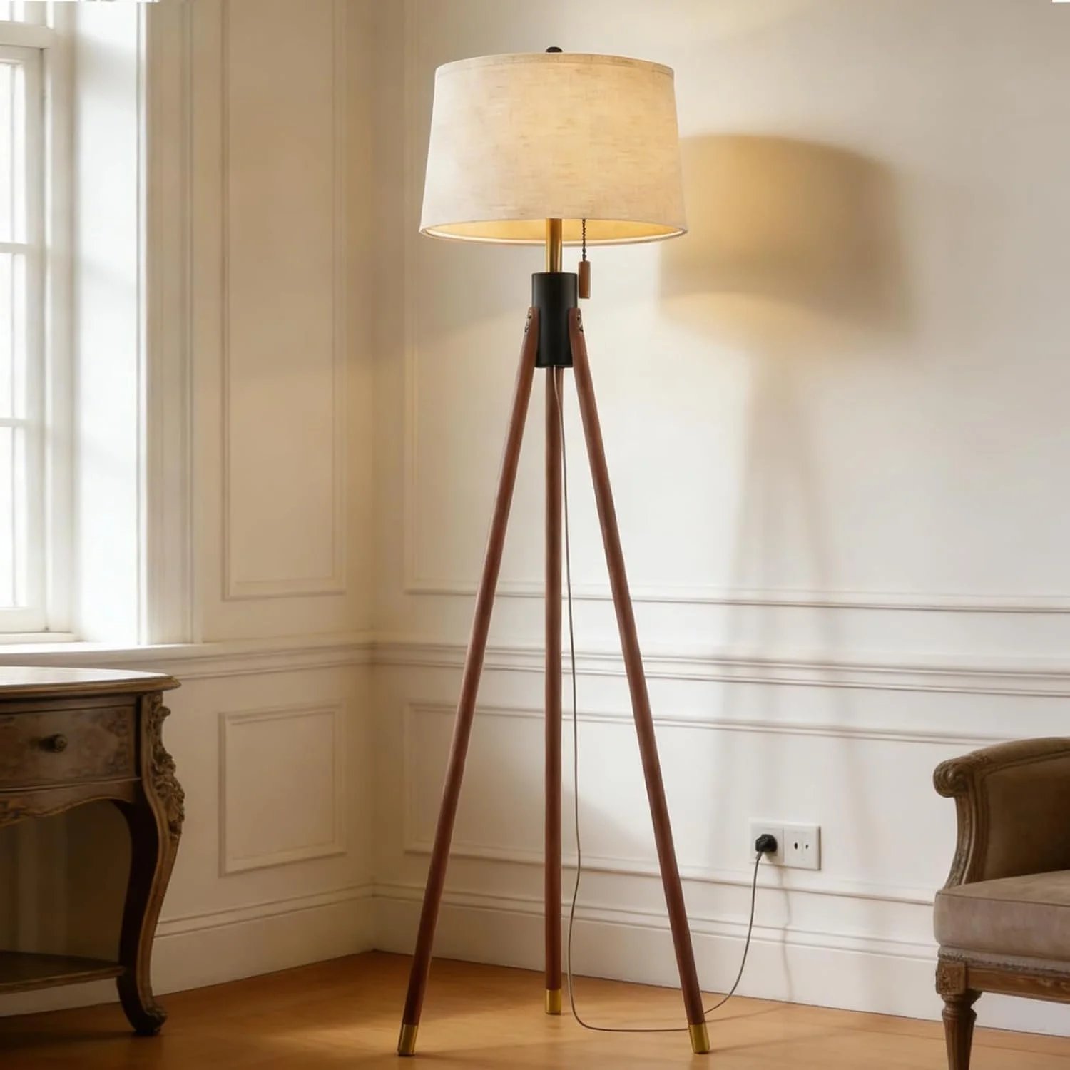 A tall floor lamp with a beige lampshade, wooden tripod legs, and a pull chain switch, positioned in a room with cream-colored paneled walls, a wooden floor, a vintage-style side table, and a cushioned armchair.