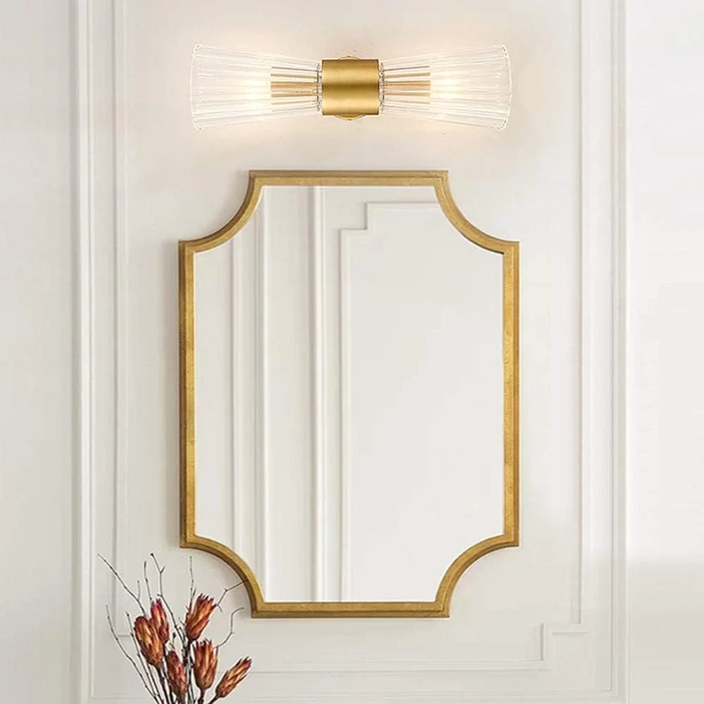 A gold-framed mirror, a modern wall sconce with glass rods, and dried flowers in a vase on a white wall.