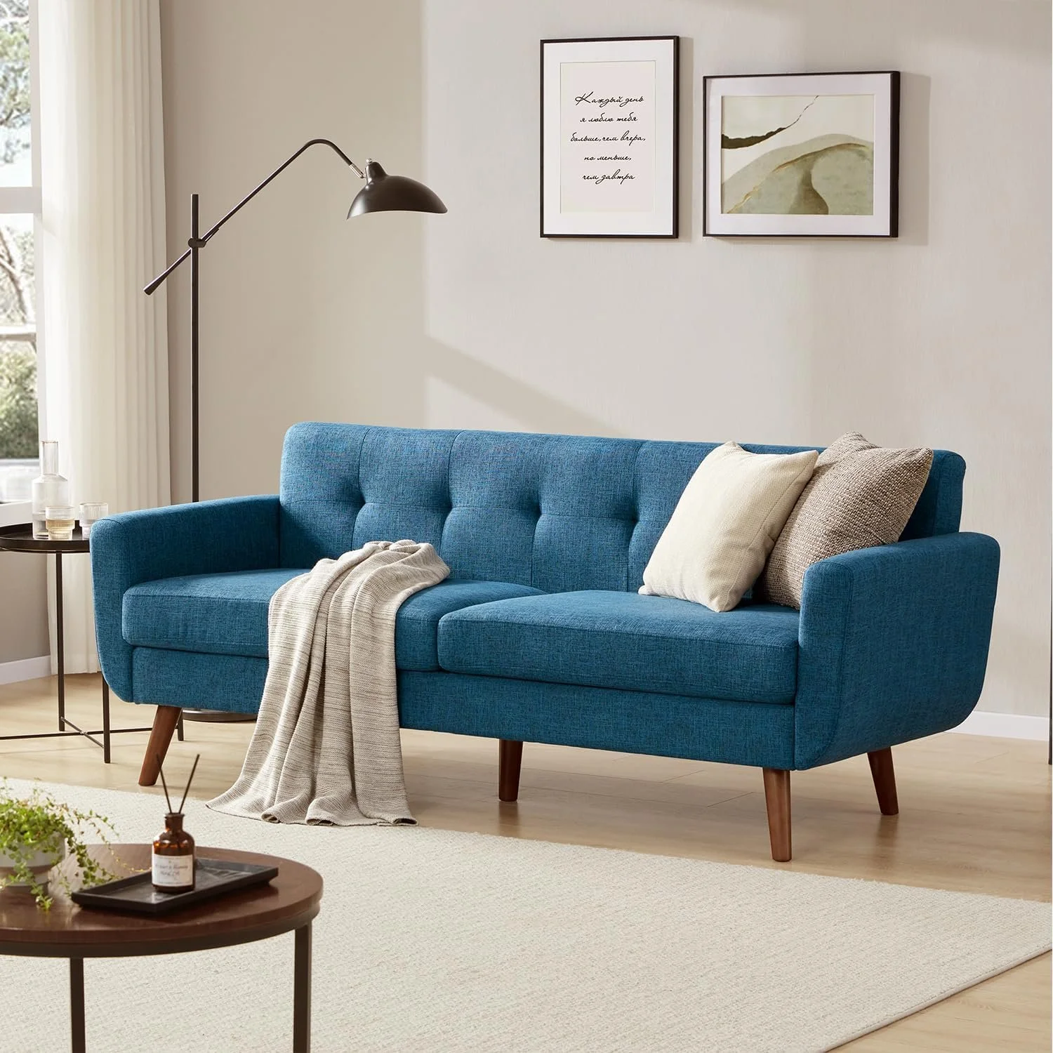 A living room with a blue danish MCM sofa, beige and brown throw pillows, a beige blanket, a black arm lamp, a small round black side table with glasses and a bottle, a beige carpet, a window with white curtains, and framed artwork on the wall.